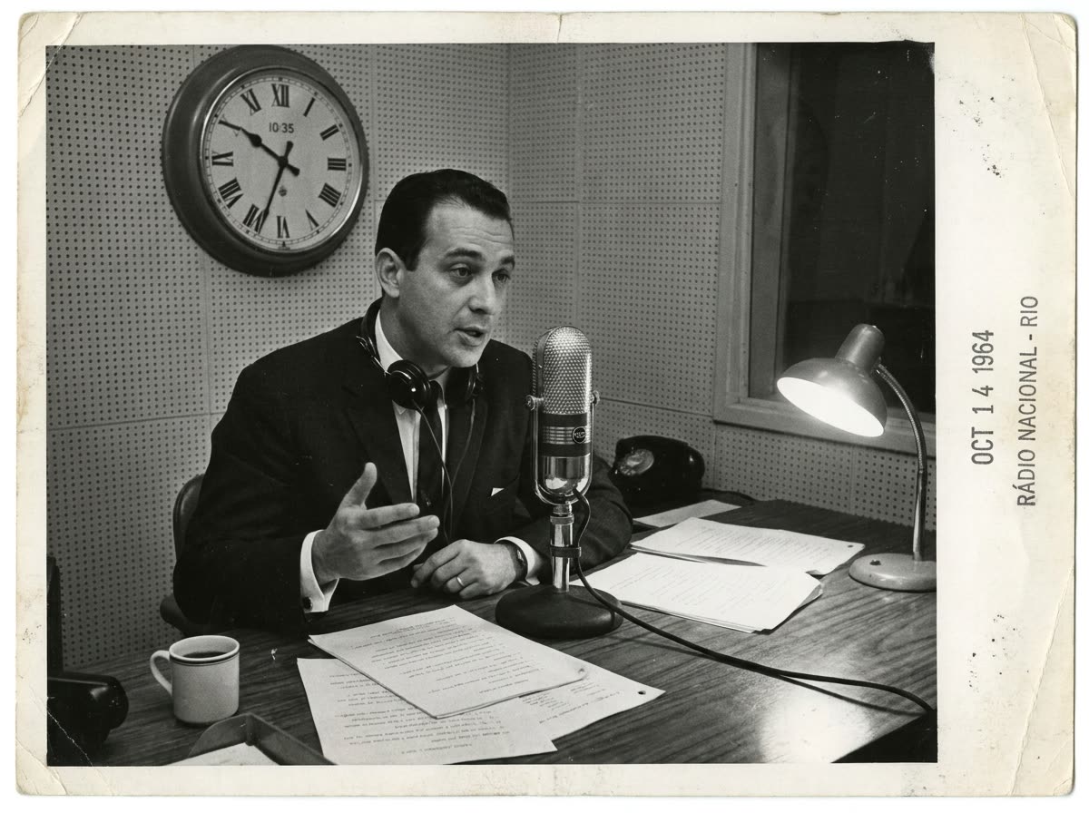 Locutor ao microfone em estúdio de rádio dos anos 1960, com relógio de parede ao fundo